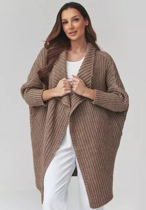 Kobieta ubrana w luźny, za duży brązowy sweter z dzianiny, nałożony na biały top i białe spodnie, trzymająca przednie brzegi swetra.