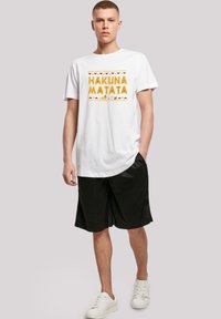 Bílý bavlněný tričko s grafikou "HAKUNA MATATA" ve žluté a oranžové barvě, kombinované s černými šortkami a bílými teniskami. Jednoduchý design, volný střih.