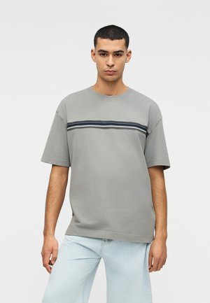 Noormees kannab halli lühikeste varrukatega t-särki, millel on üle rinna horisontaalne must-valge triip, ning helesiniseid teksapükse.