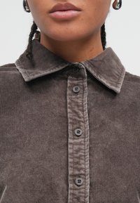 Brūna kordurojas krekls ar apkakli, sešām pogām un smalku ribotu tekstūru, uzsverot tā materiālu un dizaina detaļas.