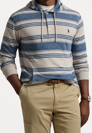 Man wearing blue and beige striped hoodie, beige pants with braided brown belt, and a wristwatch, hands in pockets.