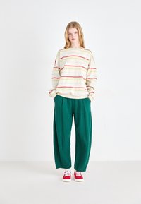 Oversized krem pulover s višeslojnim horizontalnim prugama u raznim bojama, uparen s labavim zelenim hlačama; model nosi crvene tenisice. Glatka tekstura tkanine.