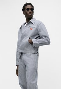 Άνδρας που φοράει σακάκι με ρίγες και αντίστοιχο παντελόνι, με το ένα χέρι στην τσέπη, φορώντας μαύρα γυαλιά ηλίου, κοιτάζοντας ελαφρώς προς το πλάι.