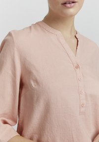 Chemise en lin rose clair avec un col en V et six boutons, dotée de manches trois-quarts et d'une coupe décontractée. Texture lisse, design minimal.