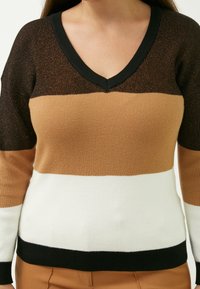 Brun, sort og hvid stribet ribbet sweater med V-udskæring og lange ærmer. Materialet har en svag glans og kontrasterende teksturer.