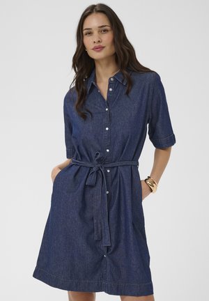 Kvinde iført en knælængde, kortærmet denimkjole med knapper foran og et bundet bælte i taljen, poserer med hænderne i lommerne mod en ensfarvet baggrund.