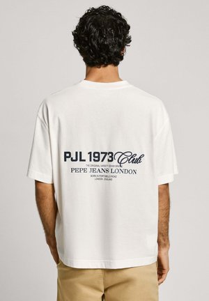 Mies, jolla on tummat kiharat hiukset ja valkoinen T-paita, jossa selässä teksti "PJL 1973 Club Pepe Jeans London", yhdistettynä beigenvärisiin housuihin.