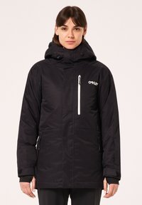 Veste noire isolée avec capuche, dotée d'une poche poitrine zippée, de poignets élastiques et d'un logo sur le côté gauche. Texture de tissu lisse.