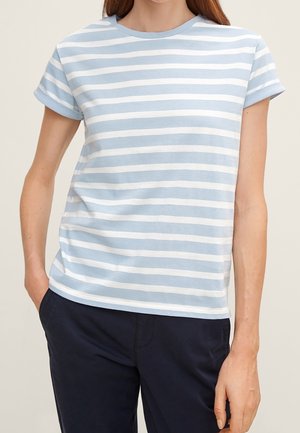 Femme portant un t-shirt à manches courtes à rayures horizontales bleu clair et blanc, et un pantalon bleu marine foncé, debout avec une main dans la poche.