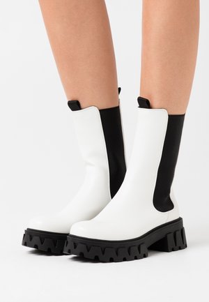 Botines blancos con paneles laterales elásticos negros, que cuentan con una suela negra gruesa y dentada. Material suave con un acabado brillante.