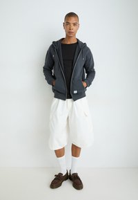 Mørk grå hettegenser med glidelås og frontlommer, svart t-skjorte, hvite cargo-shorts, brune loafers og hvite sokker mot en lys bakgrunn.