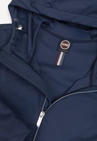 Veste à capuche bleu marine avec fermeture éclair argentée partiellement ouverte, capuche à cordon de serrage avec bouton métallique, et étiquette Colmar Originals à l'intérieur de l'encolure.