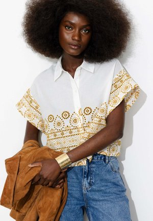 Femme avec un grand afro portant une chemise blanche à motifs floraux jaunes et un jean bleu, tenant une veste en daim marron et portant un bracelet manchette en or.