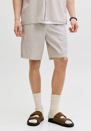 Mann iført beige og hvite stripete shorts, matchende skjorte, kremfargede sokker, brune mokkasandaletter og svart perlearmbånd.