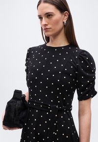 Γυναίκα που φοράει μαύρο φόρεμα με πουά σχέδιο, φουσκωτά μανίκια και ζώνη, κρατώντας μια μικρή μαύρη γούνινη τσάντα, κοιτάζοντας αριστερά με ουδέτερη έκφραση.