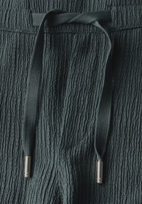Strukturierter dunkelgrüner Stoff mit elastischem Bund und passendem Kordelzug, der an den Enden mit silbernen Metallspitzen versehen ist.