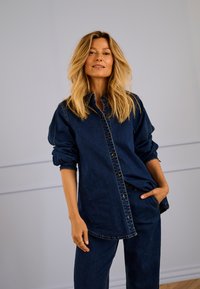 Camisa de mezclilla azul marino con cierre de botones, corte holgado, mangas remangadas y dos bolsillos en el pecho, combinada con pantalones de mezclilla a juego.