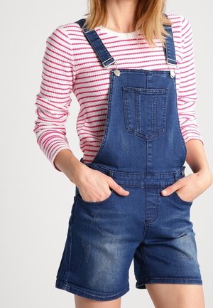 Mujer con peto corto vaquero azul y camisa blanca de manga larga con rayas horizontales rojas, manos en los bolsillos, torso visible.