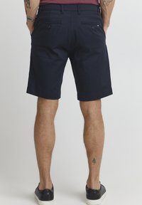 Shorts navy en tissu tissé, dotés de deux poches arrière avec des boutons et d'un ourlet retroussable. Portés avec des baskets noires, montrant des tatouages sur les jambes.