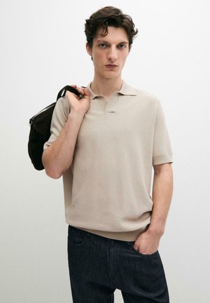 Junger Mann trägt beige strukturiertes Kurzarm-Poloshirt und dunkle Jeans, hält schwarzen Rucksackriemen über der Schulter, Hand in der Tasche.