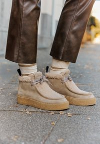 Bottes chelsea en suède marron clair avec doublure en fourrure, lacets beige et semelle en caoutchouc texturé. Portées avec des chaussettes crème et un pantalon en cuir brun ajusté.
