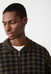 Melna un tumši zaļa vilnas jaka ar šaha rakstu, pievienots rāvējslēdzis un mīksts, teksturēts apkakle.