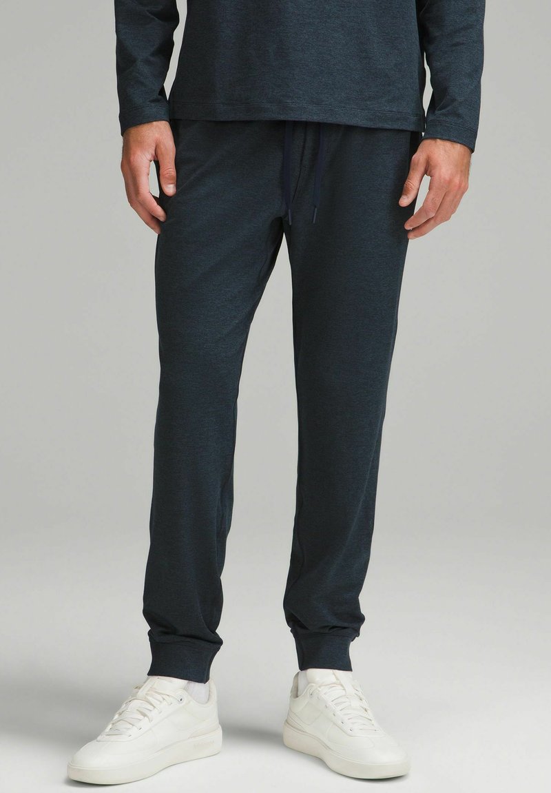 Tmavomodré jogger nohavice vyrobené z mäkkej, štruktúrovanej tkaniny, s elastickým pásom na šnúrku a zúženými manžetami. Nosí sa s bielymi teniskami.