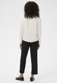 Hvid langærmet bluse med en bagfold, parret med sorte cropped bukser og sorte slip-on sko, set fra ryggen. Minimalistisk design.
