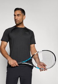 Černé krátké rukávy sportovní tričko s raglánovými rukávy, s jemným logem. Model drží tenisovou raketu s modrým akcentem.