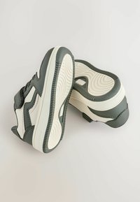Ciemnozielone buty z białymi akcentami i teksturowaną podeszwą, umieszczone na jasnym tle, podkreślające ich sportowy design.