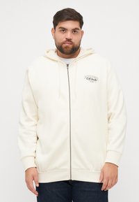 Kremfarget zip-up hettegenser laget av myk stoff, med snor i hetten og brodert logo på brystet. Ribbestrikkede mansjetter og nederkant.