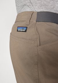 Patagonia brūnie bikses ar gludu tekstūru, melnu jostasvietu un zīmola uzlīmi ar kalnu grafiku uz aizmugurējā kabatas.