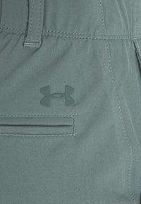 Svetle tyrkysové látkové šortky s viditelnou přední kapsou a texturovanou nášivkou loga, se švy podél pásku a kapsy.