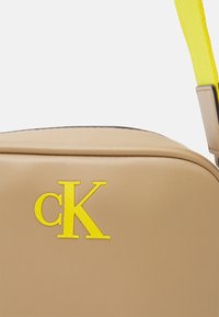 Borsa sintetica color beige con una texture liscia, caratterizzata da un prominente logo giallo "CK" e da una tracolla gialla in contrasto.