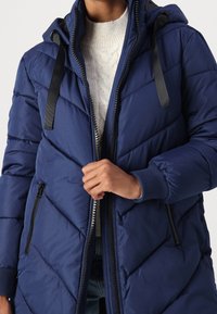 Granatowy pikowany płaszcz puchowy z przeszywanym wzorem, zamkiem błyskawicznym z przodu, kieszeniami i kapturem. Charakteryzuje się kontrastowymi czarnymi zamkami oraz ozdobnymi tasiemkami.