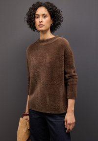 Braunmelierter, flauschiger Pullover mit hohem Kragen und Dreiviertelärmeln, kombiniert mit dunklen Jeans und einer hellen Wildledertasche.
