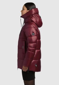 Burgundy puffer jacket with a high collar, padded design, multiple pockets, and adjustable drawstrings, featuring a circular logo patch.
