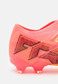 Botas de fútbol de color rosa neón con una parte superior texturizada, que presenta patrones geométricos y acentos en negro, y tacos de plástico transparente en la suela.