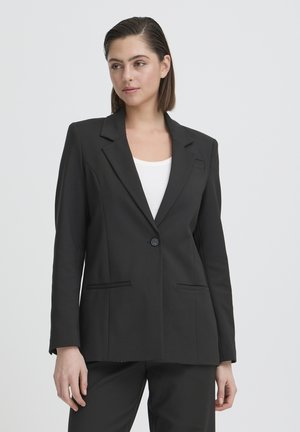 Frau mit glatten dunklen Haaren trägt einen schwarzen Blazer mit einem Knopf über einem weißen Oberteil, steht vor einem schlichten hellen Hintergrund und schaut nach rechts.