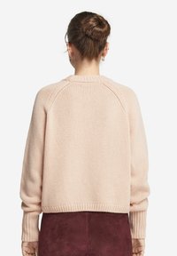Rosa gestrickter Pullover mit geripptem Saum und Bündchen. Raglanärmel und strukturiertes Gewebe. Über einem dunkelbraunen Rock getragen.