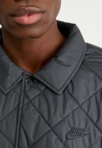 Melnā pārklāta jaka ar klasisku apkakli, kurai ir melna Nike loga izšuvums un teksturēti auduma akcenti.
