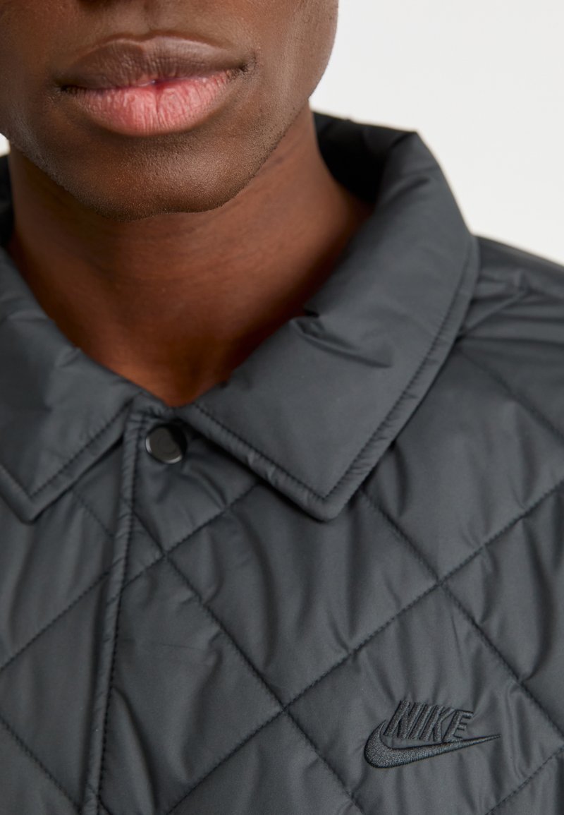Chaqueta acolchada negra con cuello clásico, que presenta bordado del logo de Nike en negro y detalles de tejido texturizado.
