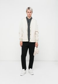 Light beige bomber jacket with front buttons, worn over a dark grey polo shirt. Black pants and white sneakers. Holding a beige cap.