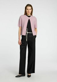 Korte roze gebreide cardigan met korte mouwen, open voorkant en gestructureerde afwerking, in combinatie met zwarte hooggetailleerde broek en zwarte platte schoenen.