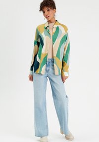 Chemise colorée à manches courtes avec des motifs ondulés verts, jaunes et bleus, assortie à un jean large bleu clair et des bottines beiges.