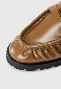 Chaussure marron brillante avec un bout arrondi, texture froncée à l'avant, et une semelle en caoutchouc noire présentant un motif de semelle contrasté.