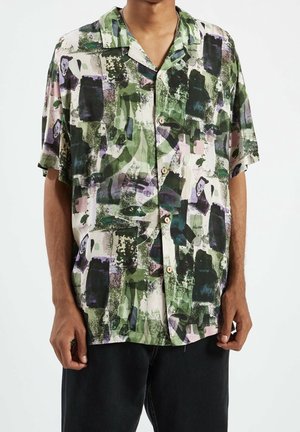 Man draagt een kort mouwen overhemdd met knopen met een groen, zwart en paars abstract patroon en zwarte broek tegen een witte achtergrond.