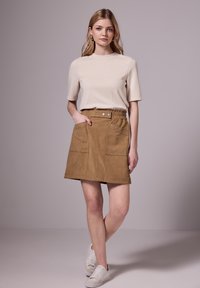 Béžový krátký rukáv top spárovaný s světle hnědou semišovou mini sukní se dvěma předními kapsami a stříbrnými ozdobami po okrajích. Jsou na sobě bílé tenisky.