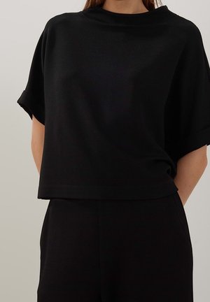 Frau trägt ein locker sitzendes schwarzes Kurzarmshirt und passende schwarze Hose vor einem schlichten hellen Hintergrund.