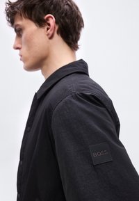 Νεαρός άνδρας με μαύρο πουκάμισο με κουμπιά και κεντημένη ετικέτα BOSS στο πάνω μέρος του μανικιού, κοιτάζοντας προς τα αριστερά μπροστά σε απλό ανοιχτό φόντο.